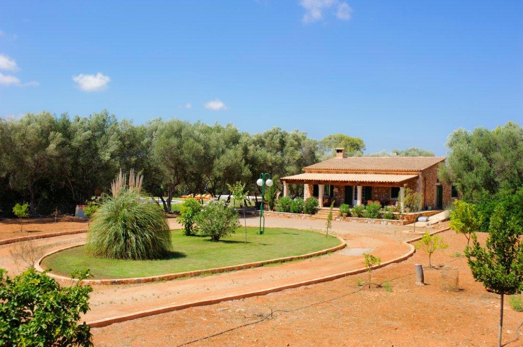 The finca seen across its garden: a circular lawn and raked-earth paths, the stone house and its pergola beyond, olive trees all around