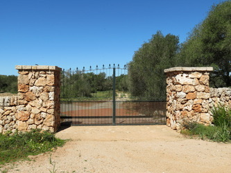 The dry-stone entrance: two rubble-stone gateposts and an iron gate opening onto the red-earth track, an olive tree beside it