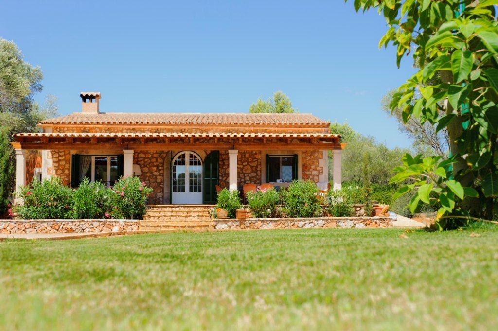 Finca Gallicant from the lawn: a long single-storey stone farmhouse with a tiled roof, a tiled pergola and an arched green door, almond trees behind under a clear blue sky