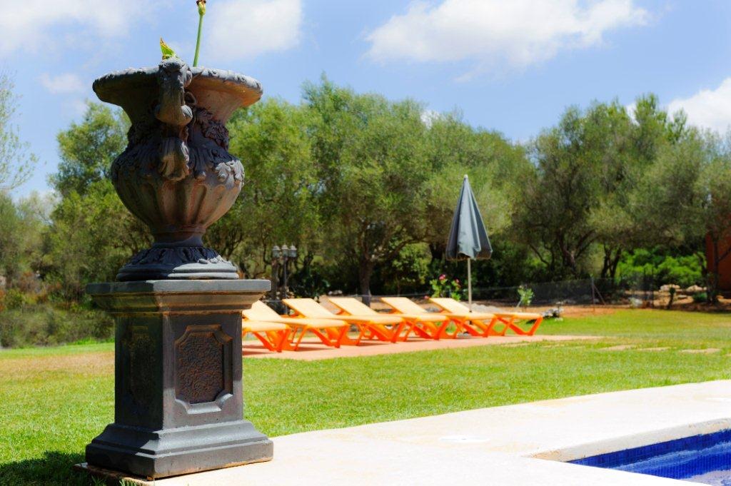 An ornamental cast urn on a plinth at the pool's edge, the loungers and lawn soft behind it