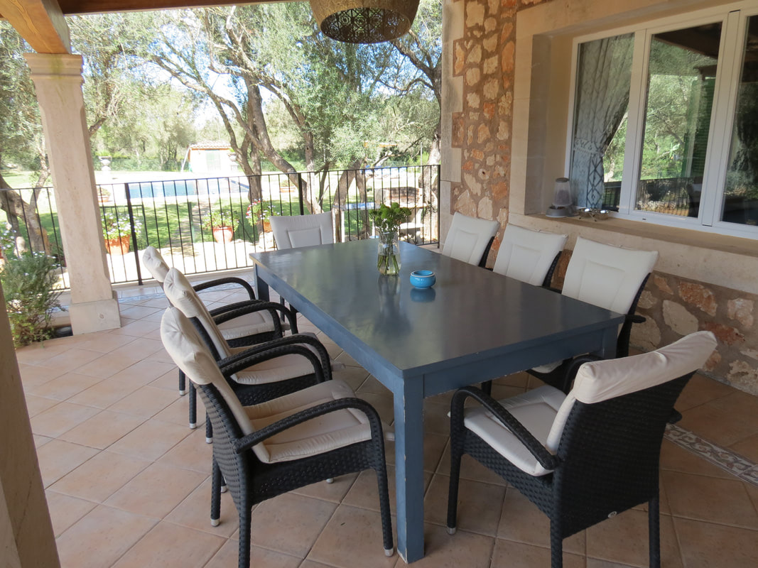The shaded terrace laid for a meal: a long table and woven chairs against the stone wall, the pool and garden visible past the railing