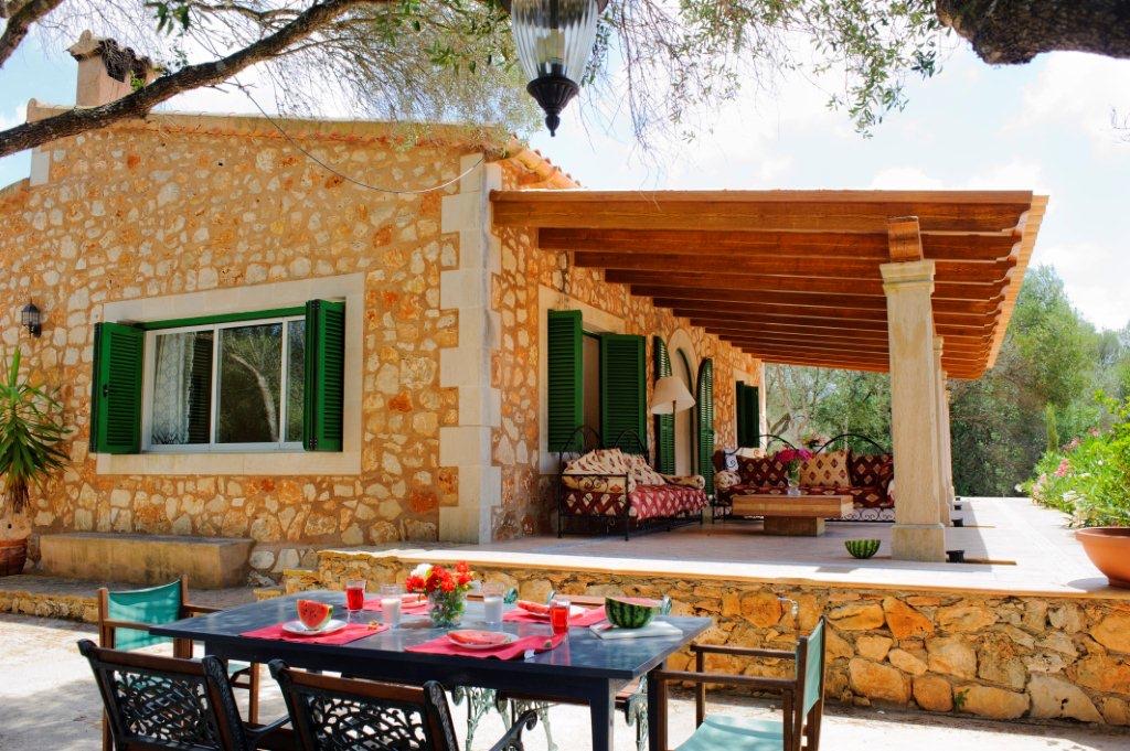The stone house and its timber pergola from the garden, a table set with watermelon and red linens in the foreground