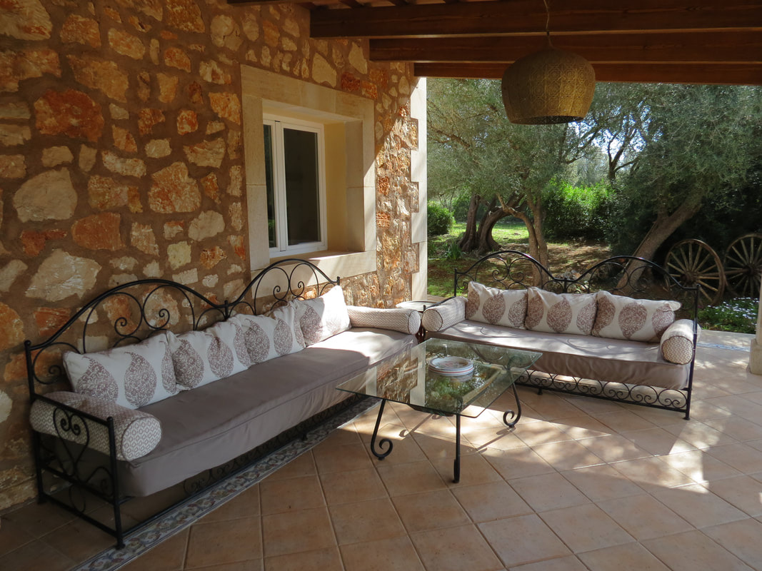 The covered stone terrace: two long wrought-iron sofas with cushions and a glass table, a woven pendant lamp, olive trees beyond